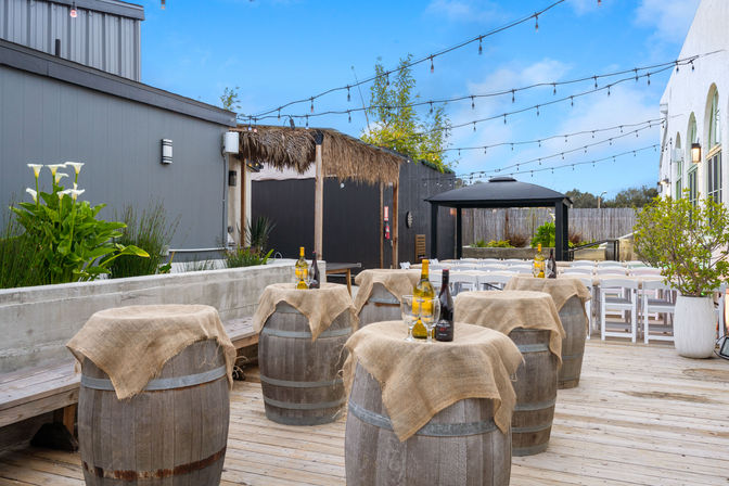 Sunny outdoor wine-tasting patio with wooden barrels draped in burlap and topped with wine bottles and glasses, string lights overhead, white folding chairs, a shaded gazebo and potted plants on a wooden deck.