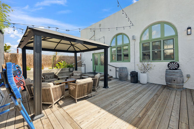Sunny outdoor wooden deck patio with black-frame covered gazebo, cushioned lounge seating, string lights, green arched windows, barrel planters and a dartboard.