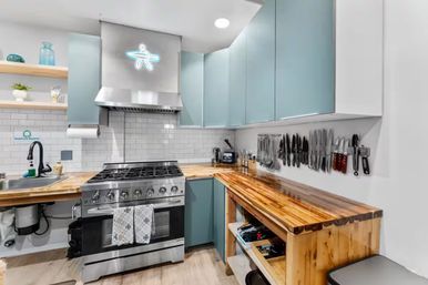 Bright modern kitchen interior with stainless steel gas range and hood, light-blue cabinets, polished butcher-block countertops, white subway-tile backsplash, magnetic knife rack and open shelving.