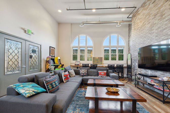 Bright loft-style apartment community lounge with arched windows, large gray sectional sofa with colorful patterned pillows, wooden coffee tables, wall-mounted TV, arcade games and study desks.