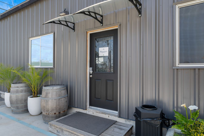Industrial gray corrugated exterior storefront entrance with a black glass‑panel door under a curved awning, wooden step and mat, two rustic barrels and potted palms, trash can and planter with white flowers.