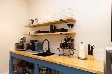 Compact modern kitchenette with light wood countertop and blue base cabinet, black sink and faucet, open wood shelves with wine glasses, plates and mugs, coffee maker, toaster oven, utensil rack, paper towel and knife block on white subway tile backsplash.