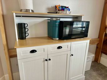 White farmhouse-style kitchenette cabinet with wood countertop holding a teal Keurig coffee maker, black microwave, stacked black mugs, paper towel roll and a small snack tray — compact kitchen corner.