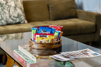 Wooden tray of assorted snack packets on a glass coffee table with brochures and a board game underneath, green sofa in a cozy living room setting