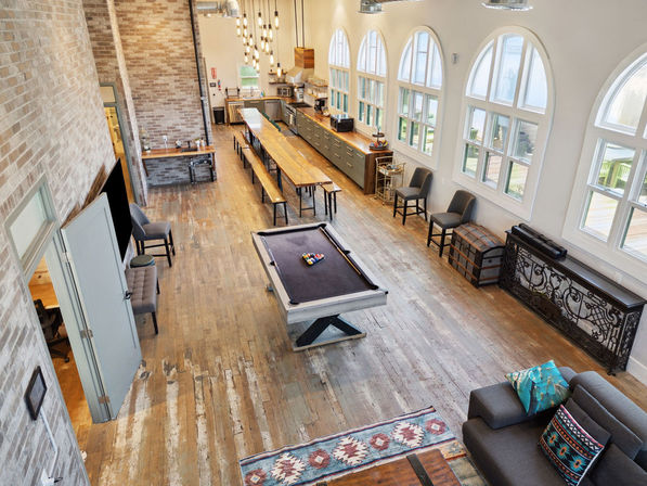 Loft-style communal room with a black-felt pool table, long wooden farmhouse dining table, arched windows, exposed brick walls, rustic hardwood floors and kitchenette seating