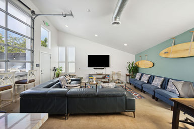 Modern coastal living room with high vaulted ceiling and large glass garage-style door, black leather sectional facing a wall-mounted TV, teal accent wall with mounted surfboards, blue bench seating, bar stools, coffee tables and indoor plants.