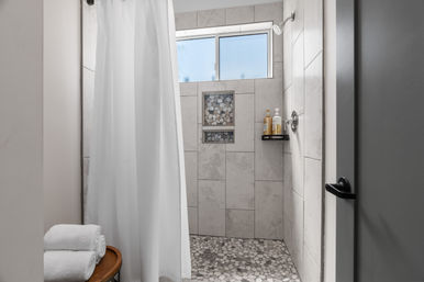 Spa-like modern walk-in shower with light stone-look wall tiles, pebble-mosaic floor, recessed niche with pebble accent, frosted window, white shower curtain, and folded towels on a wooden stool.