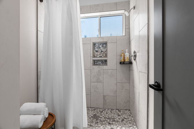 Spa-like modern walk-in shower with light stone-look wall tiles, pebble-mosaic floor, recessed niche with pebble accent, frosted window, white shower curtain, and folded towels on a wooden stool.