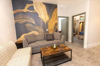 Cozy modern vacation-rental living room with a gray sofa, patterned cream loveseat, rustic wooden coffee table, and bold navy wall mural featuring oversized gold tropical leaves; doorway reveals a bunk-bed bedroom.