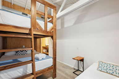 Cozy loft guest room with sturdy wooden bunk beds, single bed with patterned pillow, exposed white ceiling beams, small wooden side table, concrete floor, and neatly rolled towels by an open bathroom doorway.