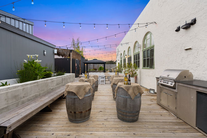 Cozy outdoor wooden patio at sunset with string lights, wine-barrel high tables draped in burlap, bench seating, potted plants, a gazebo in the distance and a stainless-steel grill.