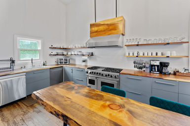Bright modern kitchen with a live-edge wood island, gray lower cabinets, wood countertops, stainless steel range and hood, open floating shelves with dishes and a coffee station.