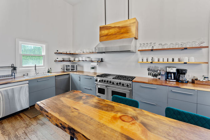Bright modern kitchen with a live-edge wood island, gray lower cabinets, wood countertops, stainless steel range and hood, open floating shelves with dishes and a coffee station.