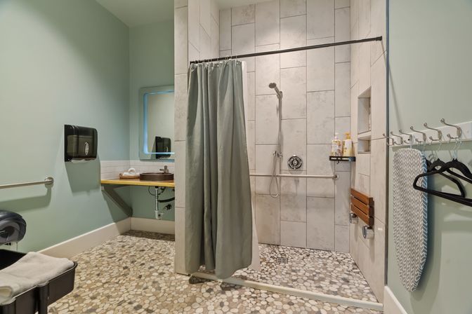 Accessible, spa-style bathroom with roll-in tiled shower, pebble-mosaic floor, green shower curtain, wall grab bars, handheld showerhead and fold-down wooden seat; floating sink with mirror, wall hooks and hanging ironing board on soft green walls.