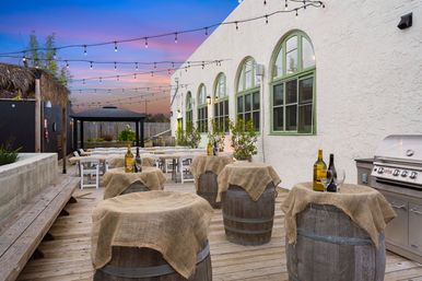 Cozy outdoor patio at sunset with string lights over a wooden deck, wine barrels draped in burlap used as cocktail tables with bottles and glasses, folding dining tables, a grill, and arched green windows on a stucco building.