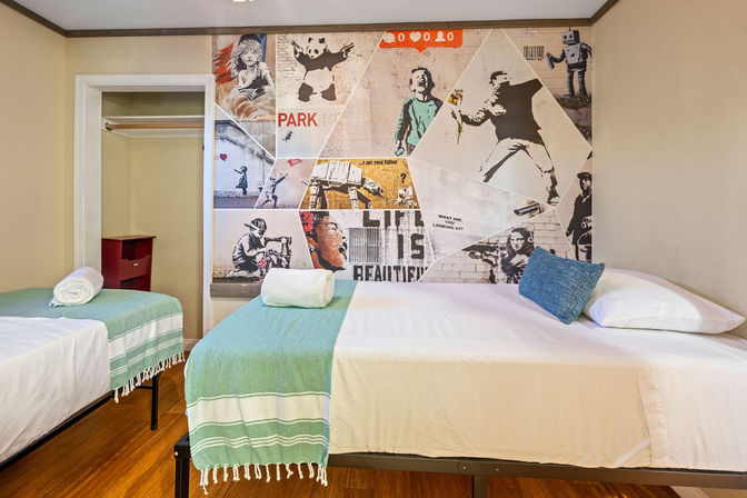 Hostel-style guest room with two single beds, teal striped throws and pillows, rolled towels, hardwood floor, open closet, and a large stenciled street-art mural on the wall.