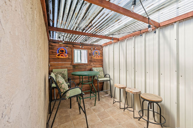 Cozy covered patio-style outdoor seating area with corrugated metal roof, wood-paneled back wall with neon icons, green bistro table and cushioned high chairs, three wooden bar stools, and hanging string lights.