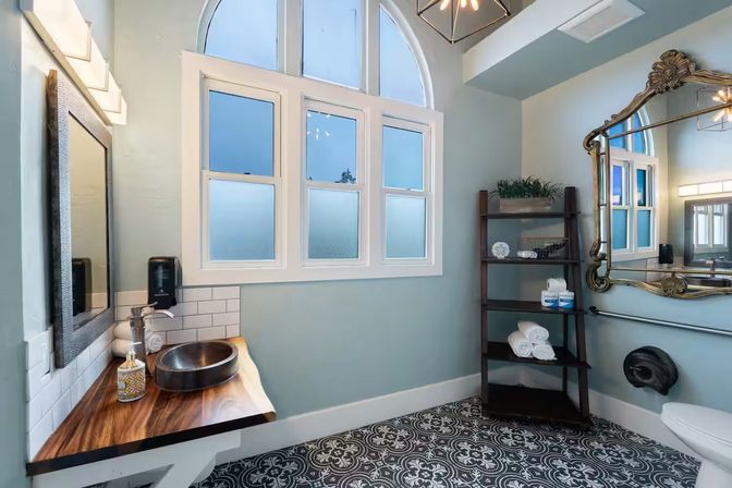 Bright modern bathroom with arched top window and soft blue walls, black-and-white patterned floor tiles, live-edge wooden floating vanity with round vessel sink, ornate gold mirror and dark open shelving stocked with rolled towels.