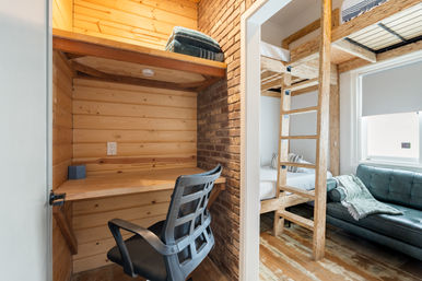 Cozy urban loft bedroom with built-in wooden study nook, black office chair, exposed brick, laddered bunk bed and green sofa by a bright window.
