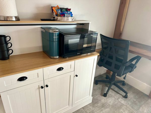 Compact kitchenette corner with teal Keurig and black microwave on a white beadboard cabinet with wooden countertop, snack tray and stacked black mugs, next to a black mesh office chair at a small desk.