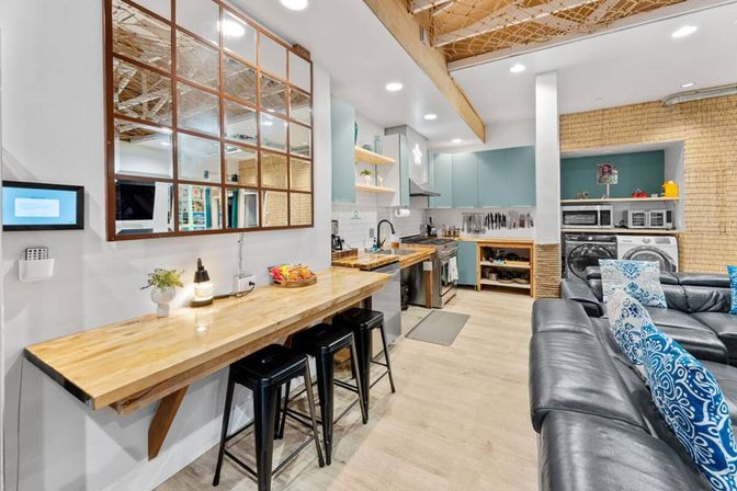 Bright open-plan basement living area with a wooden breakfast bar and three black stools, teal kitchen cabinets, stainless appliances, stacked washer and dryer in a laundry nook, and a black leather sectional with blue patterned pillows.