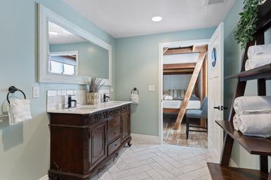 Cozy guest bathroom with carved dark wood double vanity and sinks, large white-framed mirror, mint-green walls, herringbone tile floor, towel shelf, and open door to a bunkbed bedroom with wooden beams.