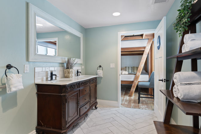 Cozy guest bathroom with carved dark wood double vanity and sinks, large white-framed mirror, mint-green walls, herringbone tile floor, towel shelf, and open door to a bunkbed bedroom with wooden beams.