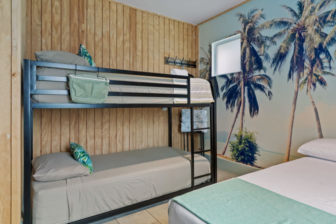 Coastal-themed bedroom with black metal bunk bed (lower mattress visible), adjacent bed, gray linens, wood-paneled wall and large palm-tree beach mural for a tropical vibe.