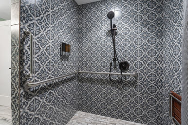 Modern walk-in shower with gray patterned tiles, black handheld showerhead, stainless steel grab bars, recessed soap niche and wooden bench — stylish accessible bathroom design.