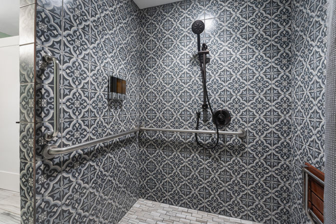 Modern walk-in shower with gray patterned tiles, black handheld showerhead, stainless steel grab bars, recessed soap niche and wooden bench — stylish accessible bathroom design.