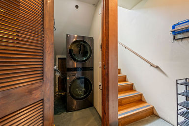 Stacked front-loading washer and dryer tucked into a compact laundry closet beneath wooden stairs, with a louvered closet door, concrete floor, wood steps and handrail, and metal shelving nearby.