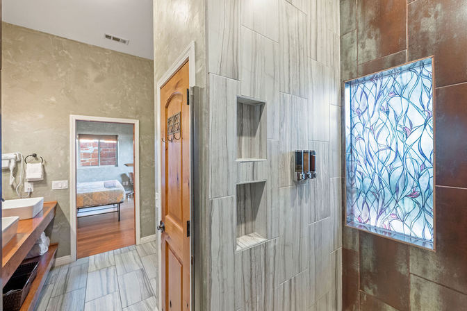 Bright modern bathroom with wooden door, marble-look tiled walk-in shower with recessed shelves and stained-glass window, wooden vanity with vessel sinks and view into bedroom