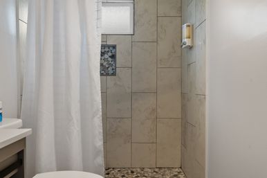 Cozy residential shower with a white curtain, frosted window, stone-look rectangular wall tiles, pebble-tile floor, recessed pebble niche and wall-mounted soap dispenser.