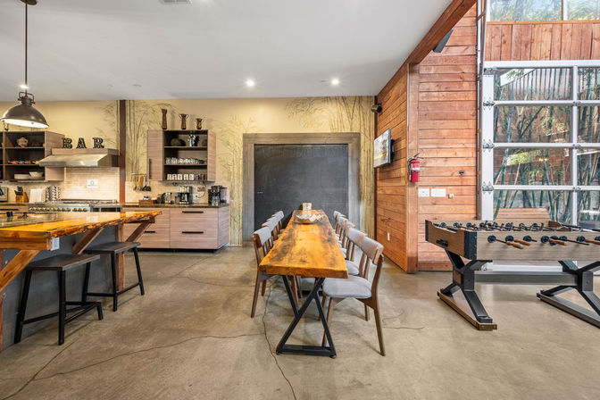 Open-plan industrial-style kitchen and communal dining area featuring a live-edge wooden table with upholstered chairs, breakfast bar with stools, foosball table, concrete floor, wood-paneled wall and large glass garage-style door bringing in natural light.