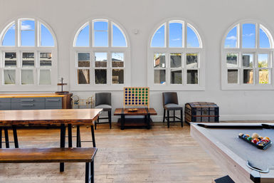 Bright, sunlit game room interior with a row of arched windows, hardwood floors, a rustic wooden communal table and bench, a pool table with racked billiard balls, two chairs flanking a small table with a four-in-a-row peg game, and a vintage trunk — ready for games.