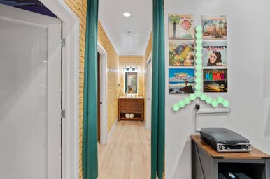 Cozy coastal-themed apartment hallway with yellow brick-pattern walls and light wood floors; teal curtains frame a mid-century vanity at the end, while vintage Hawaiian vinyl covers, mint-green hex LED panels, and a record player sit on a console.