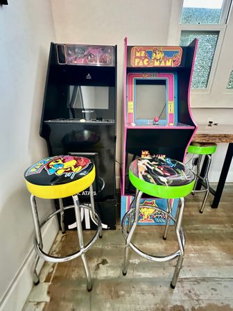 Two retro arcade machines — a black Atari cabinet and a pink Ms. Pac‑Man unit — paired with colorful Pac‑Man themed chrome bar stools on a worn wooden floor in a home game room.