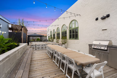 Outdoor wooden-deck courtyard with long banquet table and white folding chairs under string lights, stucco wall with arched green windows and a stainless grill at sunset.