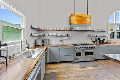 Bright modern farmhouse kitchen with live-edge wood countertops, stainless double-oven range and wooden hood, open shelves of white dishes, sink under a window and rustic hardwood floors.