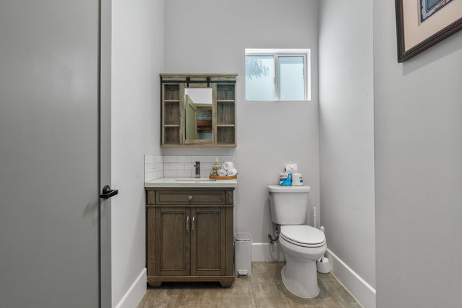 Cozy modern small bathroom interior with wood vanity and white countertop, subway tile backsplash, rustic mirrored medicine cabinet, white toilet beneath a frosted high window, gray walls and tile floor.