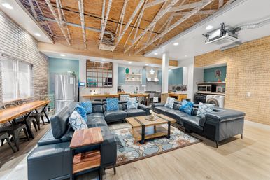 Open-plan industrial-coastal loft living area with a wraparound navy leather sectional, patterned rug, wooden coffee table, long dining table, stainless steel fridge, island kitchen, exposed wood ceiling and brick accent wall.
