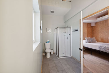 Bright modern bathroom with glass walk-in shower, white toilet, tiled floor and window, opening to a wood‑paneled bedroom with a bed
