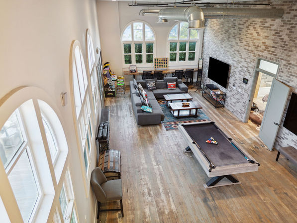 Bright industrial loft game room with arched windows, exposed ductwork and brick accent wall, sectional sofa, coffee tables, wall TV and a gray pool table on wide-plank wood floors