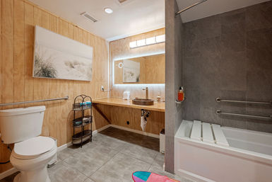 Bright accessible bathroom with light wood-paneled walls, floating wooden vanity and round vessel sink under an LED mirror; white toilet, gray-tiled tub with grab bars and fold-down seat, metal storage rack and coastal wall art.