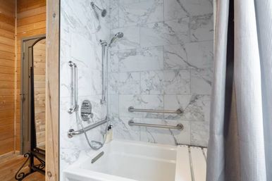 Marble-tiled bathtub and shower with dual showerheads and handheld, stainless steel grab bars, gray shower curtain, and wood-paneled wall with full-length mirror.