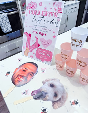 Nashville bachelorette party display on a kitchen island: pink “last rodeo” sign with cowboy boots and disco ball artwork, stacked blush cups with gold script, and playful face-and-dog photo props on sticks