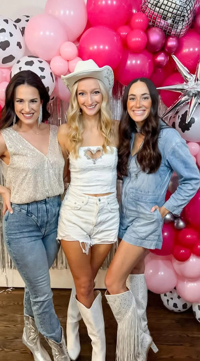 Three smiling women in cowgirl outfits — white cowboy hat, denim and fringe boots — posing at an indoor pink and cow-print balloon arch photo backdrop.