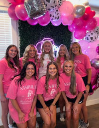 Eight women wearing matching pink Nashville t-shirts pose smiling under a hot-pink and cow-print balloon arch with metallic balloons, neon sign and greenery backdrop for a bachelorette celebration