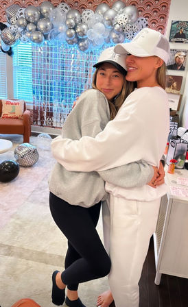 Two friends in baseball caps hugging at a cozy living-room celebration, wearing sweatshirts and leggings with silver-and-black balloon garland, disco-ball decor and a metallic photo backdrop.