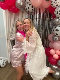 Two smiling women hugging at an indoor party, wearing satin pajamas and a sheer white robe with feather boas, posed in front of a silver fringe backdrop and a pink, silver and cow-print balloon arch.